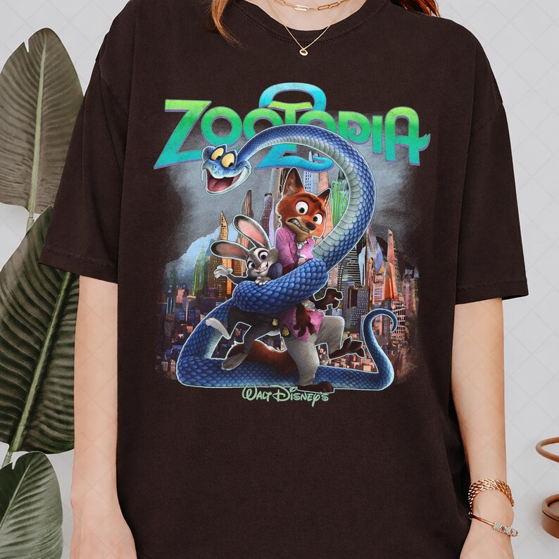 May include: A dark brown t-shirt with a vibrant graphic from the film Zootopia. The design showcases the film's title, a blue snake, and the characters Judy Hopps and Nick Wilde. The Walt Disney's logo is also visible.