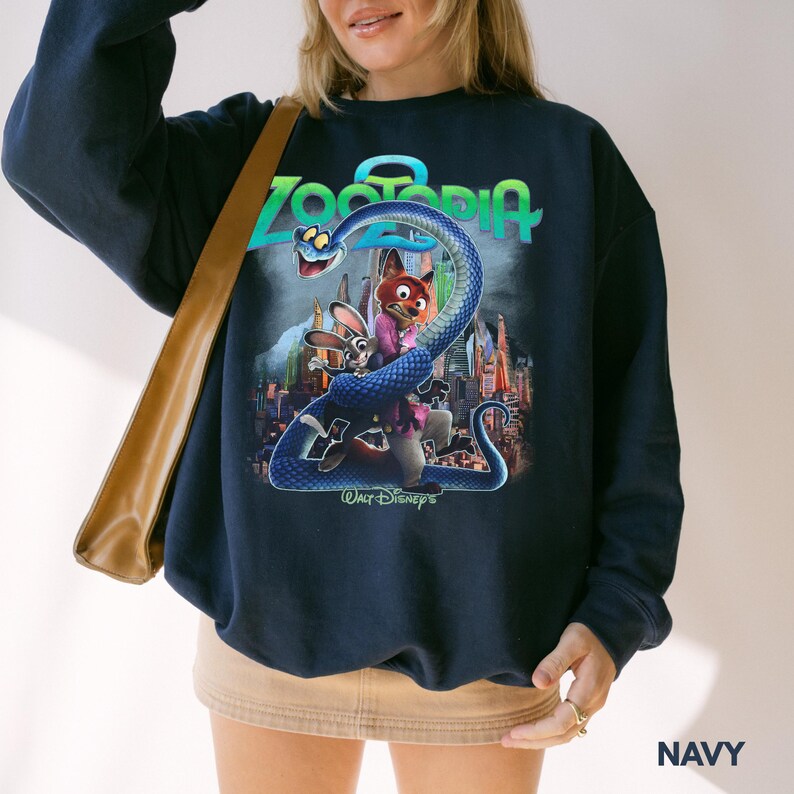 May include: Navy blue sweatshirt with a vibrant "Zootopia" graphic. The design showcases the film's title in teal, a blue snake, and the characters Nick Wilde and Judy Hopps. The words "Walt Disney's" are printed at the bottom of the design.