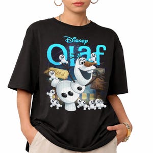May include: Black t-shirt featuring the Disney character Olaf from Frozen. The design includes Olaf, small snowmen, and the word "Olaf" in blue, with the Disney logo above. The background has a colorful, illustrated scene.