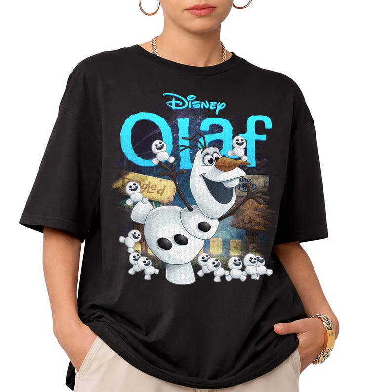May include: Black t-shirt featuring the Disney character Olaf from Frozen. The design includes Olaf, small snowmen, and the word "Olaf" in blue, with the Disney logo above. The background has a colorful, illustrated scene.