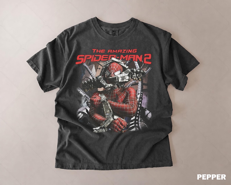May include: A dark gray t-shirt featuring a graphic from "The Amazing Spider-Man 2". The design shows Spider-Man in his costume with mechanical arms. The text "THE AMAZING SPIDER-MAN 2" is printed in red across the top.