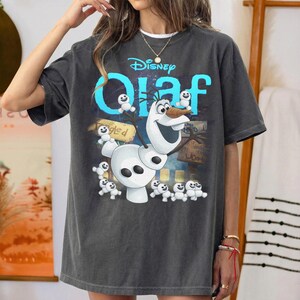 May include: Dark gray t-shirt featuring the Disney character Olaf from Frozen. The design includes Olaf, small snowmen, and the word "Olaf" in blue. The Disney logo is at the top. The shirt has a relaxed fit.