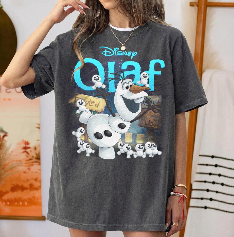 May include: Dark gray t-shirt featuring the Disney character Olaf from Frozen. The design includes Olaf, small snowmen, and the word "Olaf" in blue. The Disney logo is at the top. The shirt has a relaxed fit.