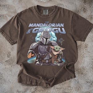 Może przedstawiać: Brązowa koszulka z grafiką The Mandalorian i Grogu z Gwiezdnych Wojen. Projekt zawiera tekst "The Mandalorian & Grogu" w kolorze niebieskim i białym, z ilustracjami postaci pośrodku.
