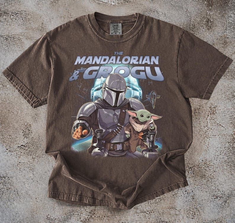 Może przedstawiać: Brązowa koszulka z grafiką The Mandalorian i Grogu z Gwiezdnych Wojen. Projekt zawiera tekst "The Mandalorian & Grogu" w kolorze niebieskim i białym, z ilustracjami postaci pośrodku.
