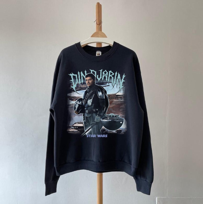 May include: Black sweatshirt featuring a graphic of Din Djarin from Star Wars, holding his helmet, with Grogu in a pod. The text "DIN DJARIN" is above the image, and "STAR WARS" is below.
