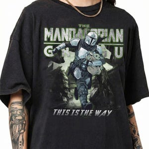 Puede incluir: Camiseta gris oscuro con un dise&ntilde;o de The Mandalorian y Grogu. El dise&ntilde;o incluye el texto "THE MANDALORIAN" en verde y blanco, y "THIS IS THE WAY" en blanco.