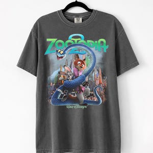 May include: Dark grey t-shirt with a Zootopia movie graphic. The design features the film's title, a blue snake, and characters Judy Hopps and Nick Wilde. The t-shirt is displayed on a black hanger.