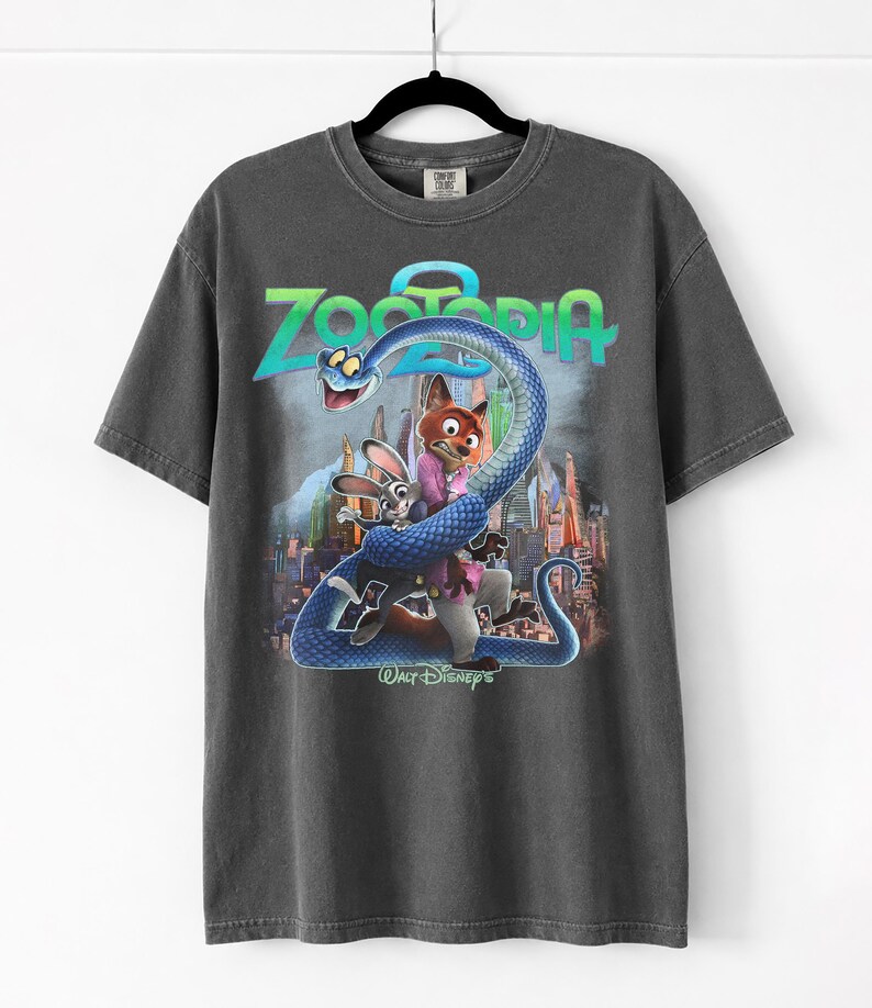 May include: Dark grey t-shirt with a Zootopia movie graphic. The design features the film's title, a blue snake, and characters Judy Hopps and Nick Wilde. The t-shirt is displayed on a black hanger.
