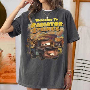 Puede incluir: Camiseta gris oscuro con un gr&aacute;fico de Mate de la pel&iacute;cula Cars, con el texto "Welcome To Radiator Springs". El dise&ntilde;o incluye un paisaje des&eacute;rtico y las palabras "Tow Mater".