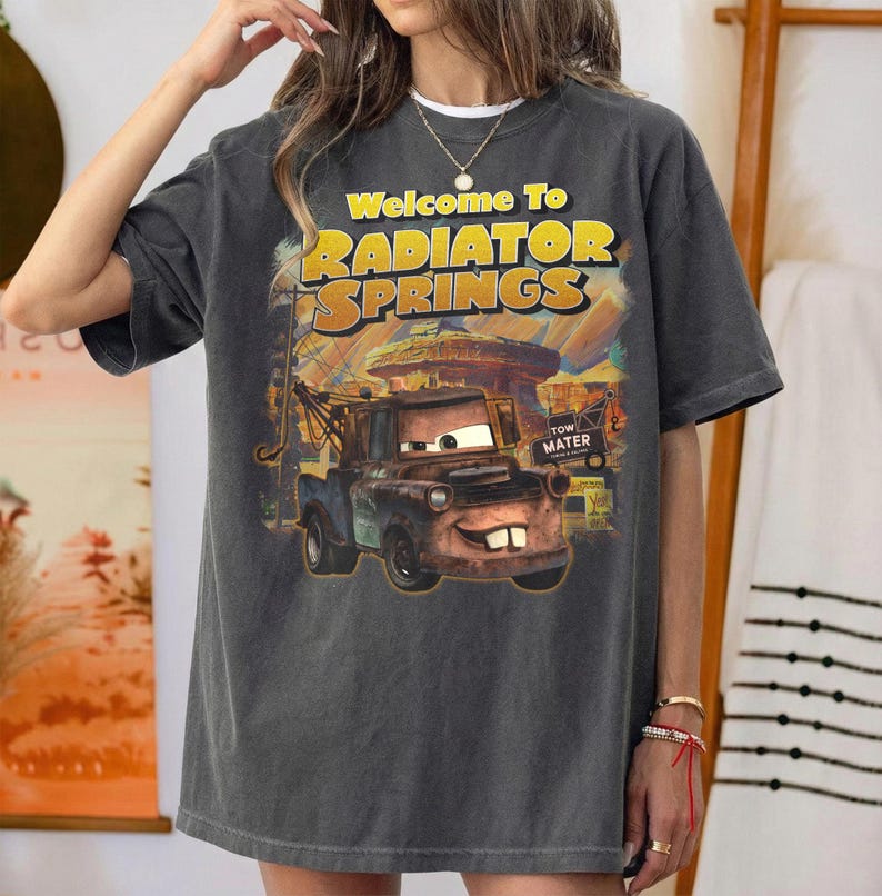 Puede incluir: Camiseta gris oscuro con un gr&aacute;fico de Mate de la pel&iacute;cula Cars, con el texto "Welcome To Radiator Springs". El dise&ntilde;o incluye un paisaje des&eacute;rtico y las palabras "Tow Mater".