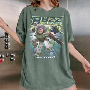 Op de afbeelding: Groen t-shirt met een afbeelding van Buzz Lightyear en een alien uit de film "Lightyear". Het ontwerp bevat de woorden "Buzz Lightyear" in geel en wit. Het shirt heeft een ronde hals en korte mouwen.