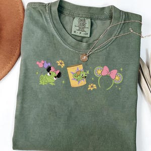 Peut inclure: Un t-shirt à manches courtes vert olive avec une impression graphique représentant un caméléon vert portant un nœud rose, une lanterne jaune avec un caméléon et des oreilles vertes avec un nœud rose. Le t-shirt porte une étiquette Comfort Colors.