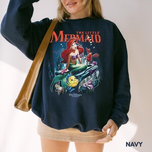 May include: Navy blue sweatshirt featuring a graphic of Ariel from Disney's "The Little Mermaid." The design includes Ariel, Flounder, Sebastian, and other characters. The text "THE LITTLE MERMAID" is displayed above the characters, with "Walt Disney CLASSIC" below.