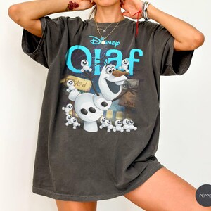 May include: Dark gray t-shirt featuring the Disney character Olaf from Frozen. The shirt has a large graphic of Olaf with the word "Olaf" in turquoise and white. The shirt is oversized and has short sleeves.