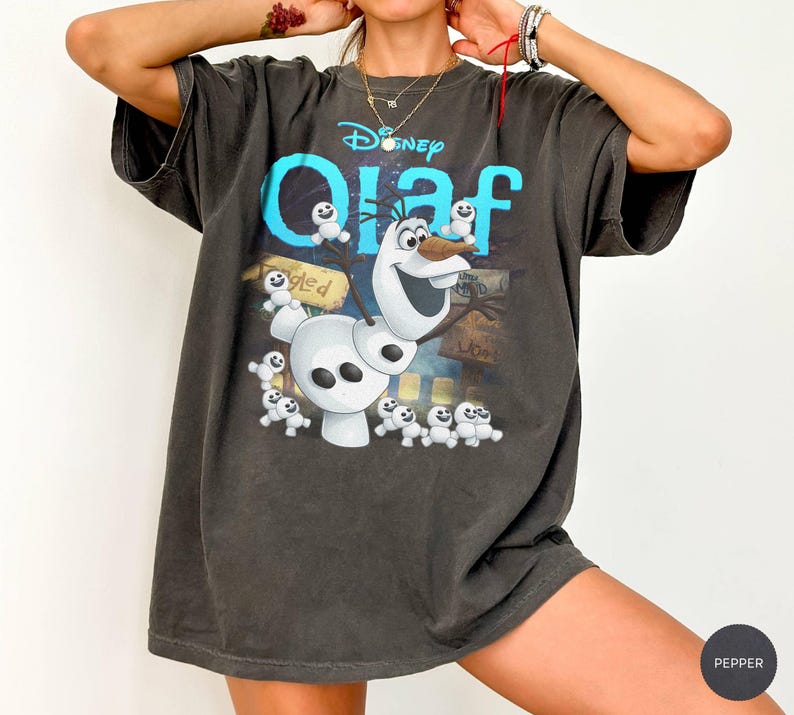 May include: Dark gray t-shirt featuring the Disney character Olaf from Frozen. The shirt has a large graphic of Olaf with the word "Olaf" in turquoise and white. The shirt is oversized and has short sleeves.