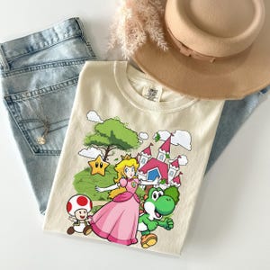 May include: A cream-colored t-shirt featuring a graphic print of Princess Peach, Yoshi, and Toad in front of a castle and a green tree. The shirt is paired with light blue denim jeans, a tan wide-brimmed hat, and a delicate silver necklace.