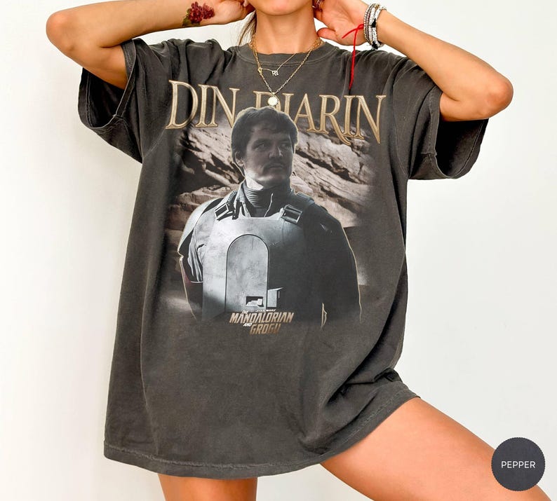 May include: Dark gray t-shirt featuring a graphic of Din Djarin from The Mandalorian. The text "DIN DJARIN" is arched above the image, with "MANDALORIAN GROGU" below. The shirt has a vintage, distressed look.