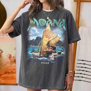 Pu&ograve; includere: T-shirt grigio scuro con una grafica colorata del film d'animazione "Moana". Il design include il titolo del film in turchese, una barca con personaggi e la parola "PIXAR" in basso. La maglietta ha una vestibilit&agrave; ampia.