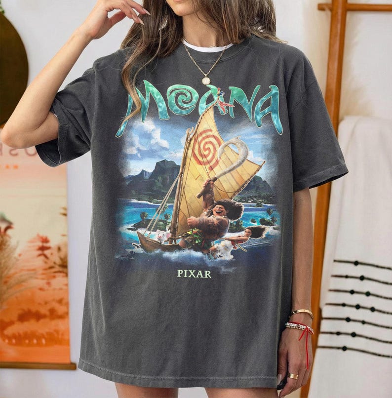 Pu&ograve; includere: T-shirt grigio scuro con una grafica colorata del film d'animazione "Moana". Il design include il titolo del film in turchese, una barca con personaggi e la parola "PIXAR" in basso. La maglietta ha una vestibilit&agrave; ampia.