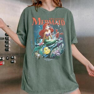 May include: Green t-shirt featuring a graphic of Ariel from Disney's The Little Mermaid. The design includes Ariel, Flounder, Sebastian, and other characters. The text "The Little Mermaid" and "Walt Disney Classic" are also visible.