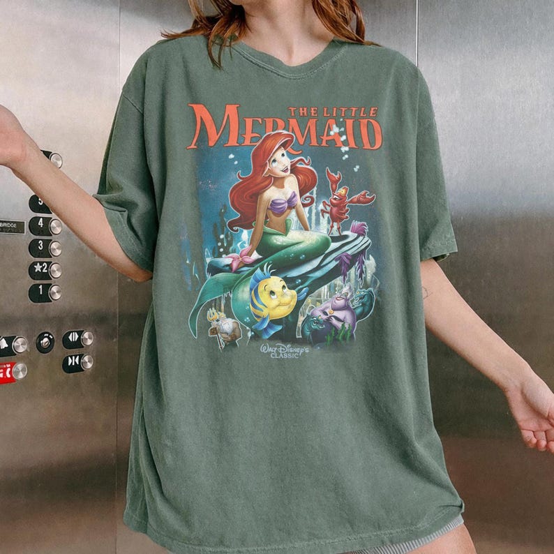May include: Green t-shirt featuring a graphic of Ariel from Disney's The Little Mermaid. The design includes Ariel, Flounder, Sebastian, and other characters. The text "The Little Mermaid" and "Walt Disney Classic" are also visible.