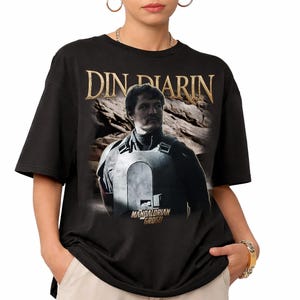May include: Black t-shirt featuring a portrait of Din Djarin from The Mandalorian. The image includes the text "DIN DJARIN" in gold lettering above the character and "MANDALORIAN GROGU" below.