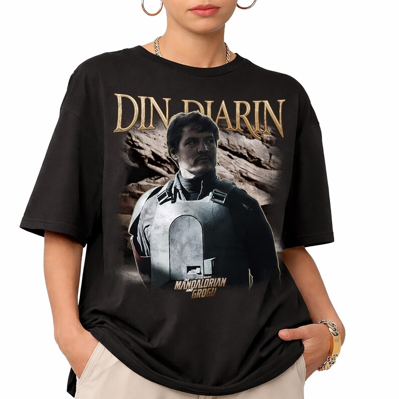 May include: Black t-shirt featuring a portrait of Din Djarin from The Mandalorian. The image includes the text "DIN DJARIN" in gold lettering above the character and "MANDALORIAN GROGU" below.