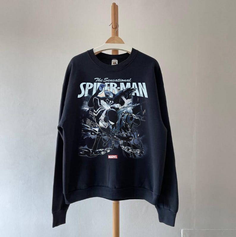 May include: Black sweatshirt featuring a graphic of Spider-Man with the text "The Sensational Spider-Man" in white and blue. The Marvel logo is at the bottom. The sweatshirt is hanging on a wooden hanger.