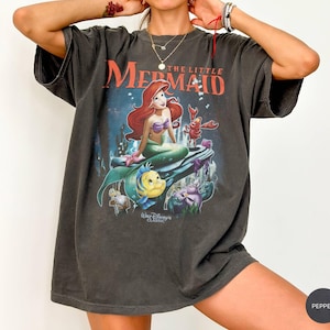 May include: Dark gray t-shirt featuring a graphic of Ariel from Disney's The Little Mermaid. The image shows Ariel with red hair, a purple top, and a green tail, along with Flounder and Sebastian. The text "The Little Mermaid" is above the image.