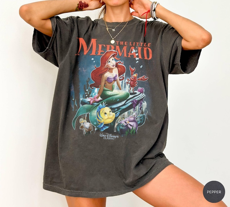May include: Dark gray t-shirt featuring a graphic of Ariel from Disney's The Little Mermaid. The image shows Ariel with red hair, a purple top, and a green tail, along with Flounder and Sebastian. The text "The Little Mermaid" is above the image.