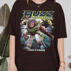 Op de afbeelding: Donkerbruin T-shirt met een afbeelding van Buzz Lightyear en een alien uit de film "Lightyear". Het ontwerp bevat de woorden "BUZZ LIGHTYEAR" in blauw en geel. De personages worden in actie afgebeeld tegen een gestileerde achtergrond.