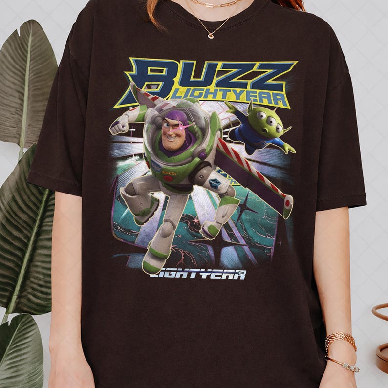Op de afbeelding: Donkerbruin T-shirt met een afbeelding van Buzz Lightyear en een alien uit de film "Lightyear". Het ontwerp bevat de woorden "BUZZ LIGHTYEAR" in blauw en geel. De personages worden in actie afgebeeld tegen een gestileerde achtergrond.