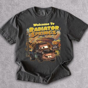 Puede incluir: Camiseta gris oscuro con un gr&aacute;fico de Mate de la pel&iacute;cula Cars, con el texto "Welcome To Radiator Springs". El dise&ntilde;o incluye una imagen de dibujos animados de Mate y el fondo de Radiator Springs.