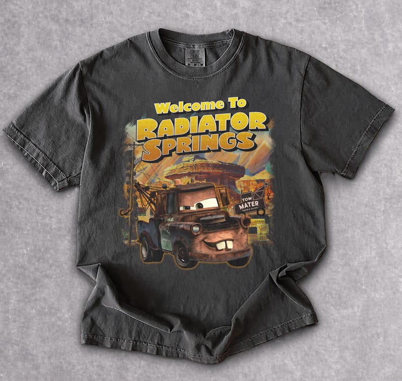 Puede incluir: Camiseta gris oscuro con un gr&aacute;fico de Mate de la pel&iacute;cula Cars, con el texto "Welcome To Radiator Springs". El dise&ntilde;o incluye una imagen de dibujos animados de Mate y el fondo de Radiator Springs.