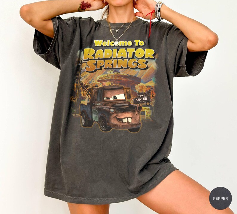 Puede incluir: Camiseta gris oscuro con un gr&aacute;fico de Mate de la pel&iacute;cula Cars, con el texto "Welcome to Radiator Springs". El dise&ntilde;o incluye un paisaje des&eacute;rtico y el nombre del personaje. La camiseta tiene mangas cortas.