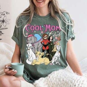 May include: A sage green t-shirt featuring the text Cool Mom Club in pink and white lettering. The graphic includes illustrations of various animated characters, including an elephant, a cat, a lion cub, a superhero, and a dalmatian dog.