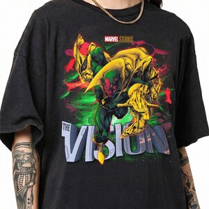 Puede incluir: Camiseta gris oscuro con un gr&aacute;fico colorido del personaje de Marvel, Vision. El dise&ntilde;o incluye el texto "THE VISION" en letras grandes y estilizadas, y el logotipo de Marvel Studios. El personaje est&aacute; representado en tonos verdes, amarillos y rojos.