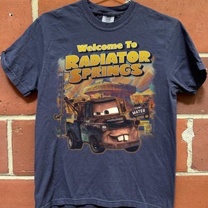 Puede incluir: Camiseta azul marino con el texto "Welcome To RADIATOR SPRINGS" en amarillo y naranja. El gr&aacute;fico representa al personaje Mater de la pel&iacute;cula Cars, con un fondo de la ciudad. La camiseta est&aacute; colgada en una percha de metal.