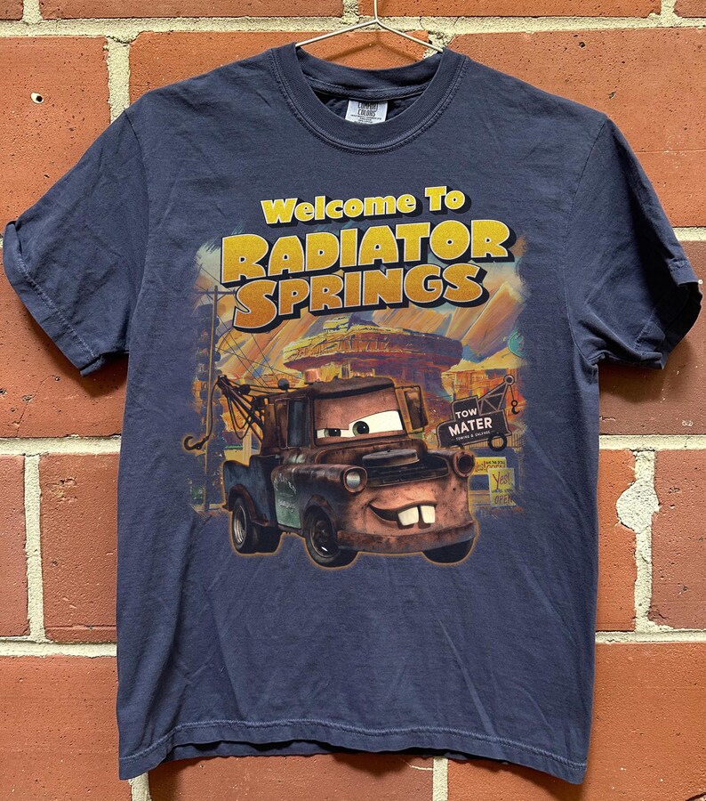 Puede incluir: Camiseta azul marino con el texto "Welcome To RADIATOR SPRINGS" en amarillo y naranja. El gr&aacute;fico representa al personaje Mater de la pel&iacute;cula Cars, con un fondo de la ciudad. La camiseta est&aacute; colgada en una percha de metal.