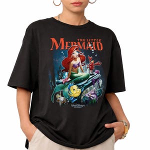 May include: Black t-shirt featuring a graphic of Ariel from Disney's The Little Mermaid. The design includes Ariel, Flounder, Sebastian, and other characters, with the text "THE LITTLE MERMAID" in orange. The shirt is a classic Disney design.