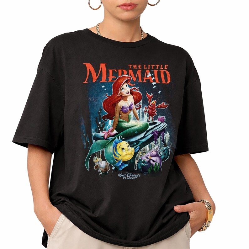 May include: Black t-shirt featuring a graphic of Ariel from Disney's The Little Mermaid. The design includes Ariel, Flounder, Sebastian, and other characters, with the text "THE LITTLE MERMAID" in orange. The shirt is a classic Disney design.