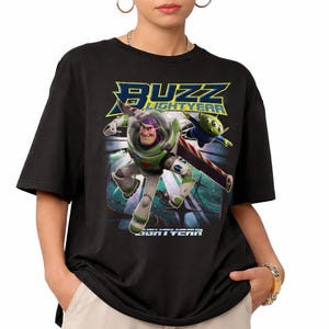 Op de afbeelding: Zwart T-shirt met een afbeelding van Buzz Lightyear en een alien uit de film Toy Story. De woorden "Buzz Lightyear" staan boven en onder de afbeelding in een blauw en wit lettertype.