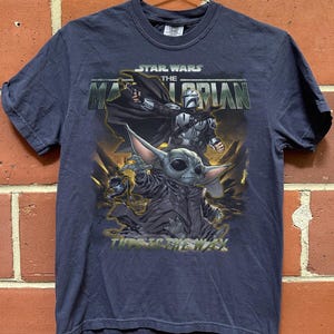 May include: Navy blue t-shirt featuring a graphic from the Star Wars series, "The Mandalorian." The design includes the Mandalorian, and Grogu, with the text "This is the Way." The shirt is short-sleeved and has a crew neck.