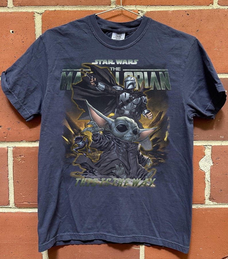 May include: Navy blue t-shirt featuring a graphic from the Star Wars series, "The Mandalorian." The design includes the Mandalorian, and Grogu, with the text "This is the Way." The shirt is short-sleeved and has a crew neck.