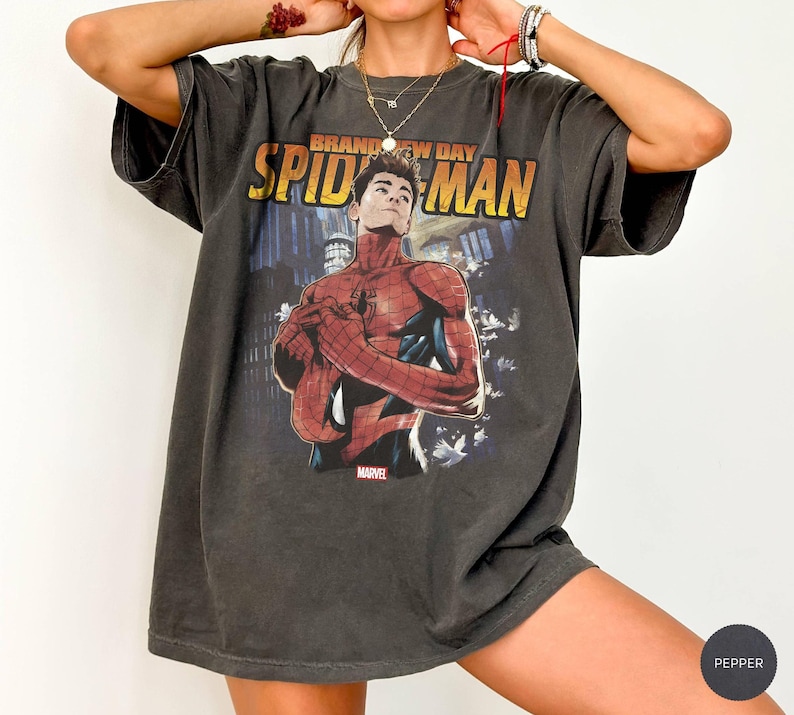 Pu&ograve; includere: T-shirt grigio scuro con grafica di Spider-Man e la scritta "Brand New Day Spider-Man" in giallo e arancione. Il logo Marvel &egrave; visibile in basso. La maglietta ha un aspetto vintage e usurato.