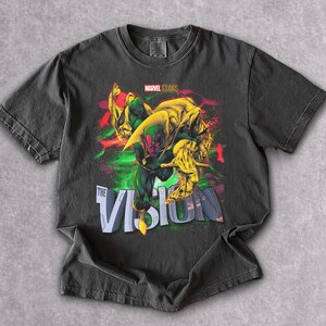 Puede incluir: Camiseta gris oscuro con un gr&aacute;fico del personaje de Marvel, Vision. El dise&ntilde;o incluye a Vision en verde y rojo, con detalles amarillos y el texto "THE VISION". Tambi&eacute;n es visible el logotipo de Marvel Studios.