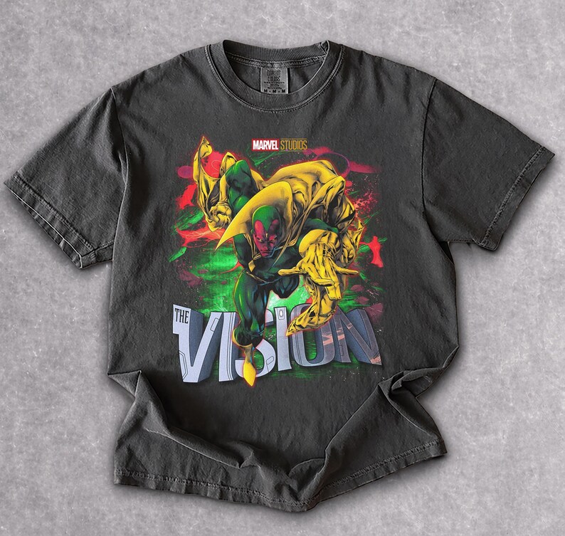 Puede incluir: Camiseta gris oscuro con un gr&aacute;fico del personaje de Marvel, Vision. El dise&ntilde;o incluye a Vision en verde y rojo, con detalles amarillos y el texto "THE VISION". Tambi&eacute;n es visible el logotipo de Marvel Studios.