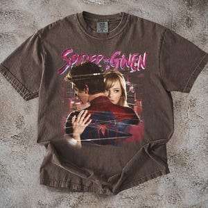 May include: Brown t-shirt with a graphic of Spider-Man and Gwen Stacy, and the text "Spider-Gwen" in pink and purple. The shirt has a vintage, distressed appearance.