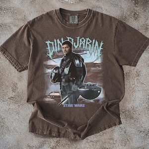 May include: Brown t-shirt featuring a graphic of Din Djarin from Star Wars, holding his helmet, with Grogu in a floating pod. The text "DIN DJARIN" is above the image, and "STAR WARS" is below.